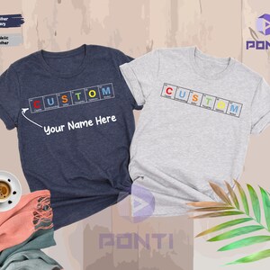 May include: Two t-shirts, one navy and one heather gray, each with the word "CUSTOM" spelled out in a periodic table-style design. The navy shirt has the text "Your Name Here" below the design. A coffee cup and a scarf are in the foreground.