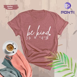 May include: Heather mauve t-shirt with the text "be kind" in white lettering and braille below. The shirt is folded and displayed on a light brown background.