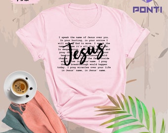 Op de afbeelding: Roze T-shirt met een zwart tekstontwerp dat luidt: "Ik spreek de naam van Jezus over je uit. In je pijn, in je verdriet zal ik God vragen om te bewegen. Ik spreek de naam omdat het alles is wat ik kan doen. In wanhoop bid ik dat genezing, vrede en kracht zullen opladen. Jezus van binnen zou een doorbraak bewerkstelligen die vandaag zou gebeuren. Ik bid voor wonderen over je leven in Jezus' naam, in Jezus' naam."