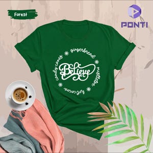 May include: Green t-shirt with a white circle design that says "Believe" surrounded by the words "hot cocoa", "candy canes", "gingerbread", and "mistletoe".