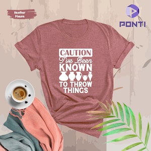 May include: Heather mauve t-shirt with white text that reads "CAUTION I've Been Known To Throw Things" with a graphic of four pottery pieces.