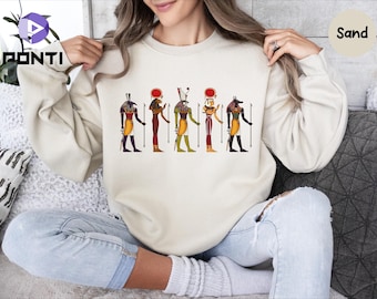 May include: Sand-colored sweatshirt featuring a colorful graphic of Egyptian deities. The design includes figures with detailed attire and staffs, in shades of yellow, red, purple, and green. The sweatshirt is a neutral tone, suitable for casual wear.