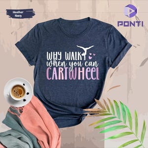 Könnte beinhalten: Ein T-Shirt in Marineblau mit dem Text "Why walk when you can cartwheel" in rosa und weißen Buchstaben. Oberhalb des Textes befindet sich eine Silhouette einer Turnerin, die einen Radschlag macht.