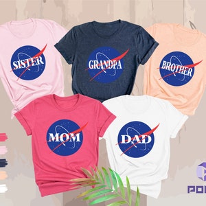 K&ouml;nnte beinhalten: F&uuml;nf T-Shirts mit einem Weltraum-Design. Die Shirts sind in Pink, Marineblau, Pfirsich, Rot und Wei&szlig;. Die Shirts haben die Worte "Schwester", "Opa", "Bruder", "Mama" und "Papa" darauf.