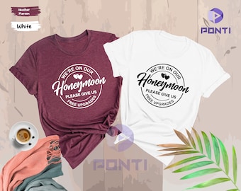 May include: Two t-shirts with a circular design that says "We're on our Honeymoon" with two hearts and the text "Please give us free upgrades". One t-shirt is white with black text and the other is maroon with white text.