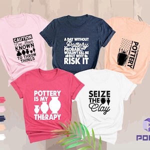 May include: Five t-shirts in various colors, each with a pottery-themed graphic. The shirts are pink, navy, peach, white, and heather raspberry. The designs include text and pottery illustrations, such as vases and clay pots.