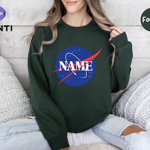 K&ouml;nnte beinhalten: Ein dunkelgr&uuml;ner Pullover mit einem blauen und roten NASA-Logo, auf dem "NAME" in roten Buchstaben steht.