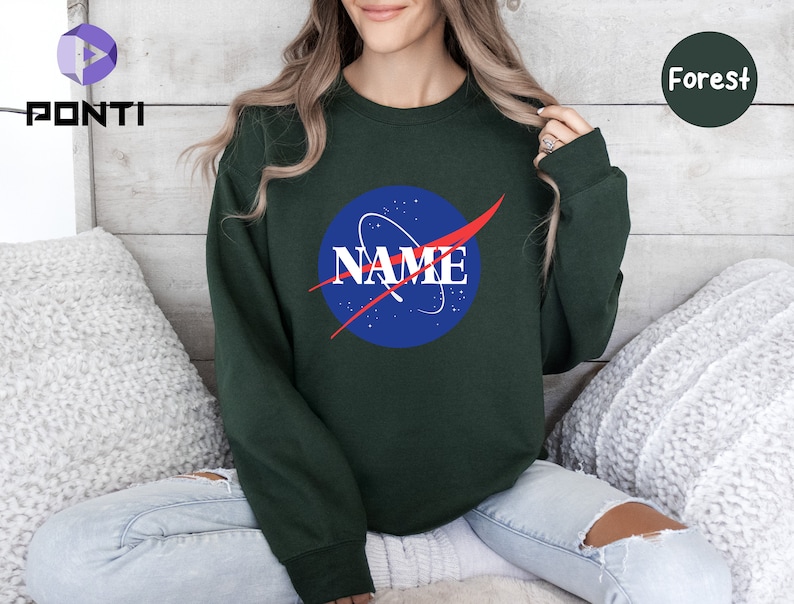 K&ouml;nnte beinhalten: Ein dunkelgr&uuml;ner Pullover mit einem blauen und roten NASA-Logo, auf dem "NAME" in roten Buchstaben steht.