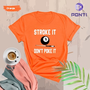 May include: Orange t-shirt with the text "STROKE IT" and a black and white 8-ball graphic. Below the graphic is the text "DON'T POKE IT".