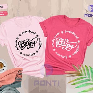 May include: Two t-shirts with a Christmas-themed design. The pink shirt has black text that says "Believe" surrounded by the words "candy canes", "gingerbread", "mistletoe", "hot cocoa", and "joy". The red shirt has white text that says "Believe" surrounded by the words "candy canes", "gingerbread", "mistletoe", "hot cocoa", and "joy".