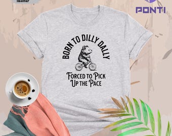 May include: A heather grey t-shirt with a black graphic of a bear riding a bicycle. The text "Born to Dilly Dally" is in a circle around the bear. Below the bear, the text reads "Forced to Pick Up the Pace".