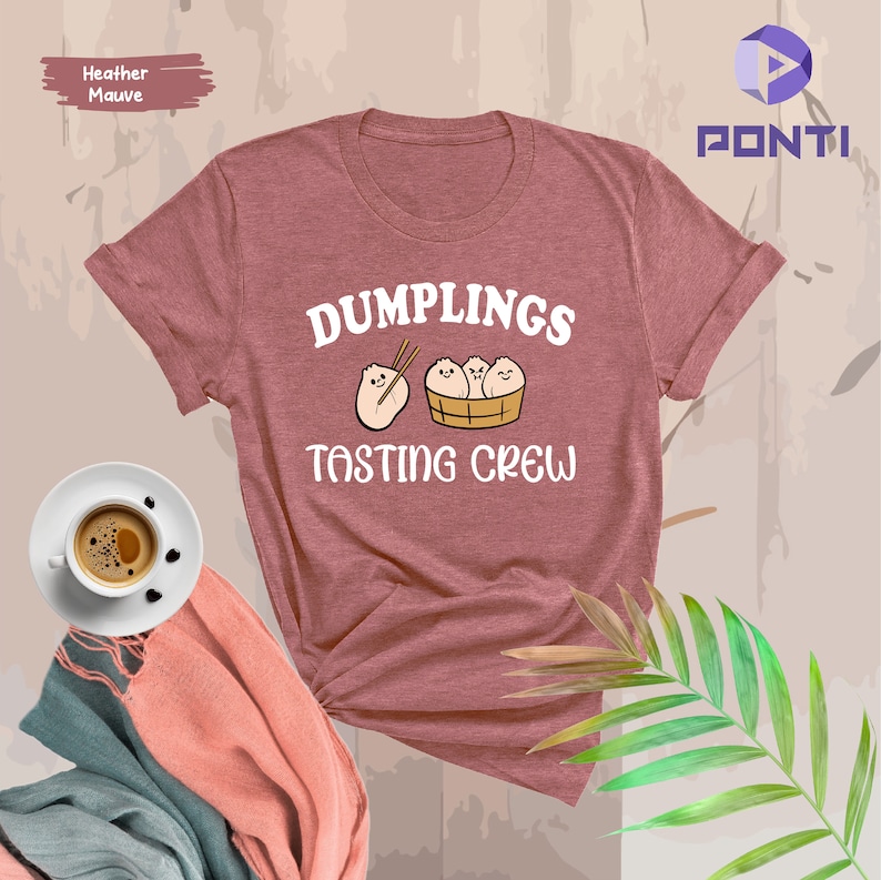 Może przedstawiać: Koszulka w kolorze heathered mauve z napisem "Dumplings Tasting Crew" w kolorze białym. Grafika przedstawia kresk&oacute;wkową ilustrację czterech pierog&oacute;w w koszyku do gotowania na parze z pałeczkami.
