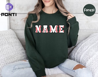 May include: A dark green sweatshirt with the word "NAME" in white block letters. The letters are filled with a baseball design, featuring red stitching. The sweatshirt is a solid color, with a round neckline.
