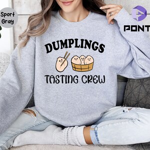 Może przedstawiać: Szara bluza z napisem "Dumplings Tasting Crew" i kresk&oacute;wkowym obrazkiem pierog&oacute;w w koszyku do gotowania na parze.