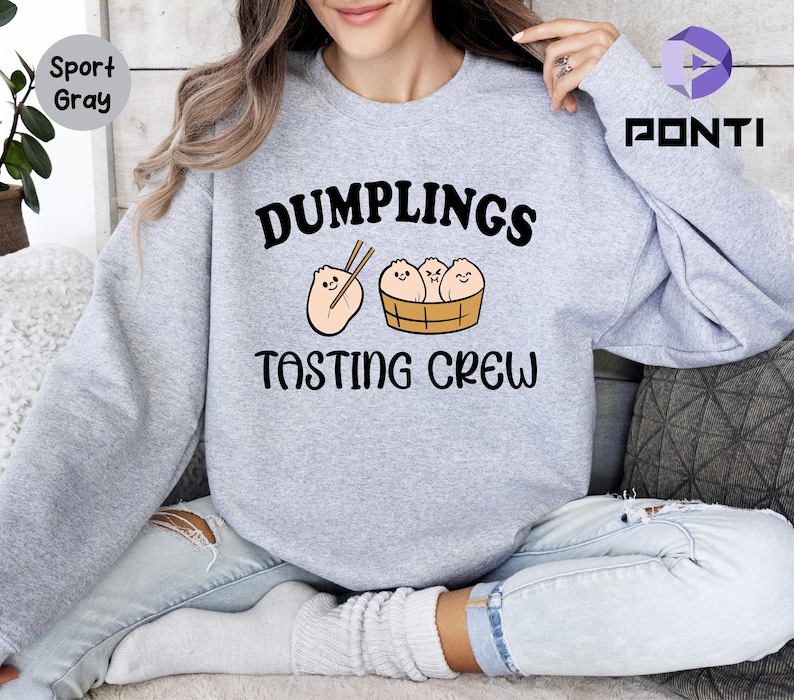 Może przedstawiać: Szara bluza z napisem "Dumplings Tasting Crew" i kresk&oacute;wkowym obrazkiem pierog&oacute;w w koszyku do gotowania na parze.