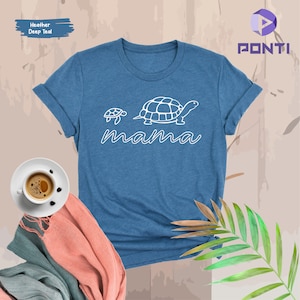 May include: A blue t-shirt with a white outline of two turtles and the word "mama" underneath. The shirt is on a light brown background with a green plant and a pink scarf.
