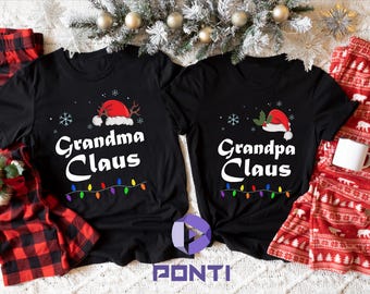 May include: Two black t-shirts with Christmas themed text and graphics. The text on the shirts reads "Grandma Claus" and "Grandpa Claus". The shirts are laid out on a red and black plaid fabric and a red and white patterned fabric.