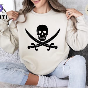 May include: A sand-colored sweatshirt featuring a black pirate skull and crossbones design. The graphic is centered on the front of the long-sleeved crew neck sweatshirt. The word "Sand" is printed on the top right of the image.
