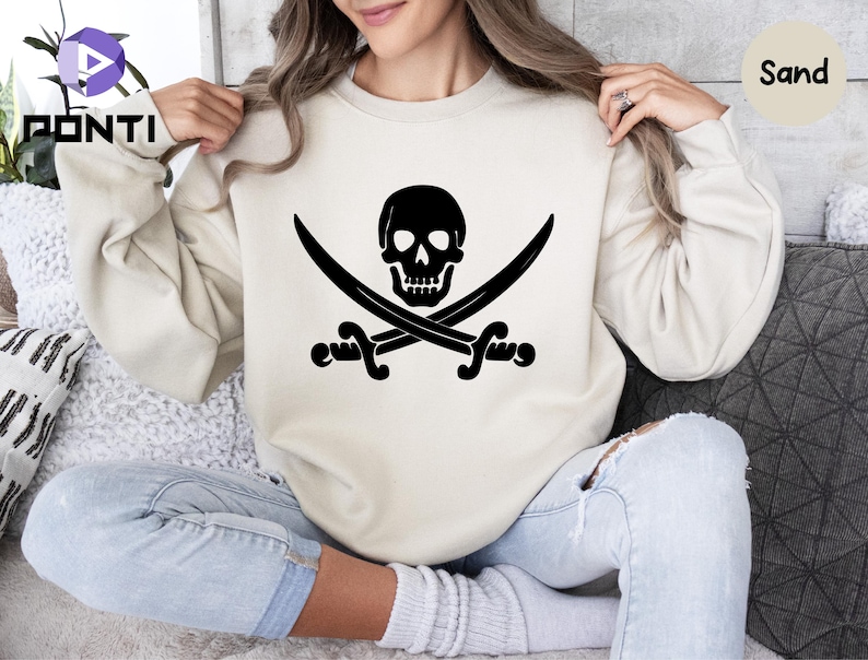 May include: A sand-colored sweatshirt featuring a black pirate skull and crossbones design. The graphic is centered on the front of the long-sleeved crew neck sweatshirt. The word "Sand" is printed on the top right of the image.