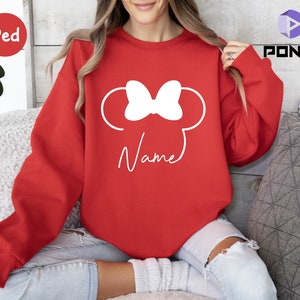 May include: A red sweatshirt featuring a white Minnie Mouse design with a bow and ears. The word "Name" is written in a script font below the design. The sweatshirt is a solid red color.