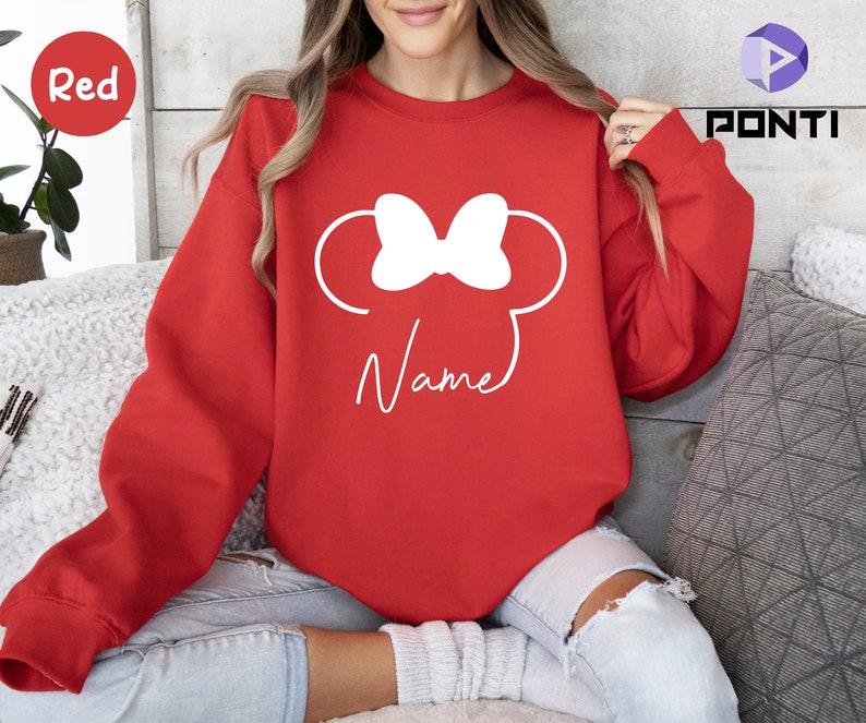 May include: A red sweatshirt featuring a white Minnie Mouse design with a bow and ears. The word "Name" is written in a script font below the design. The sweatshirt is a solid red color.