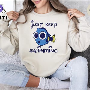 Könnte beinhalten: Ein sandfarbenes Sweatshirt mit Rundhalsausschnitt und einer Grafik eines blauen Fisches mit großen Augen und gelben Flossen. Der Text "JUST KEEP SWIMMING" ist in dunkelblauen Buchstaben über und unter dem Fisch abgebildet.