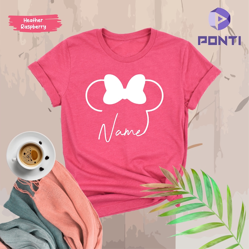 May include: A heather raspberry t-shirt featuring a white Minnie Mouse design with a bow and the word "Name" in script. The shirt has rolled-up sleeves and is displayed with a coffee cup and a scarf.