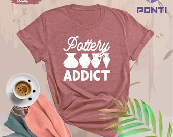 May include: Heather mauve t-shirt with the text "Pottery Addict" in white, featuring illustrations of pottery. A cup of coffee and a folded scarf are on the left. The shirt is a soft, casual style.