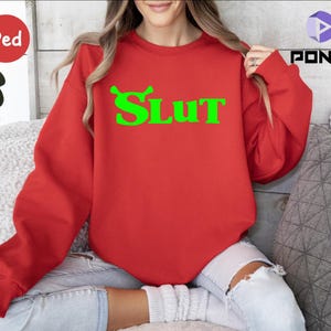 Op de afbeelding: Rood sweatshirt met een groene afbeelding van een cartoonfiguur en het woord "SLUT" in het groen.