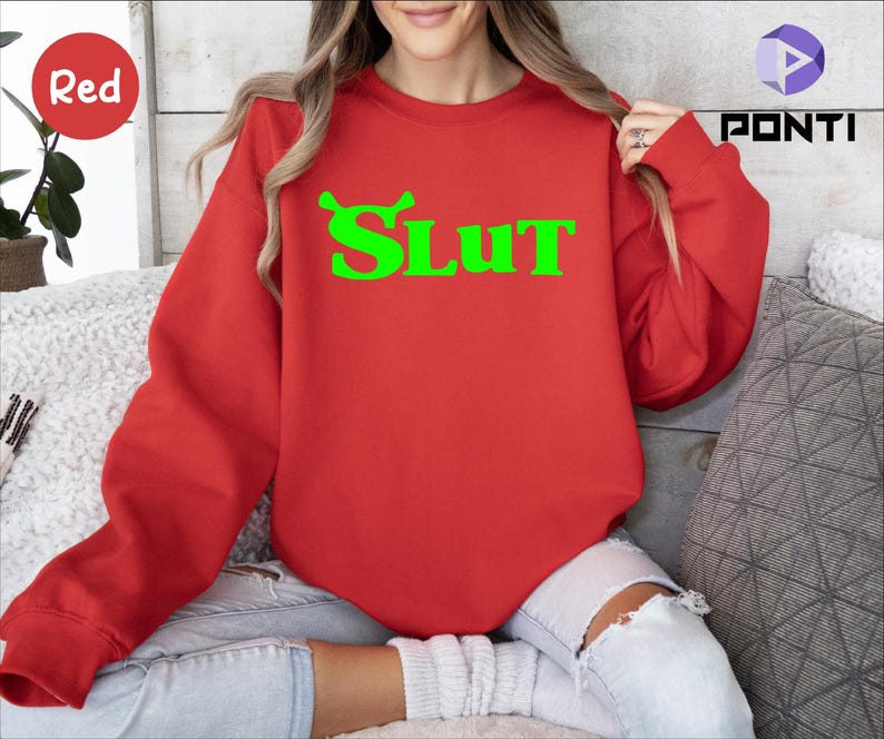 Op de afbeelding: Rood sweatshirt met een groene afbeelding van een cartoonfiguur en het woord "SLUT" in het groen.