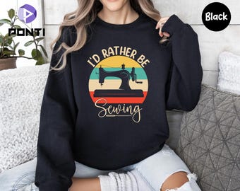 May include: Black sweatshirt with a circular graphic featuring a vintage sewing machine. The graphic includes the text "I'd Rather Be Sewing" in a retro design with yellow, teal, and red accents. The word "Sewing" is in a cursive font.