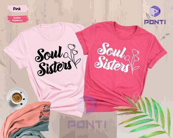 May include: Two t-shirts, one pink and one raspberry, each with the text "Soul Sisters" in a script font, accompanied by a floral design with heart-shaped blooms. The pink shirt has black text, while the raspberry shirt has white text.