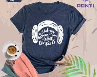 May include: Heather navy t-shirt with a white graphic of a hairstyle and the text "Well behaved women seldom defeat empires." A cup of coffee and a scarf are in the image.