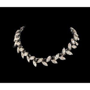 May include: A curved necklace featuring a series of clear, marquise-cut crystals set in a dark metal frame. The design consists of individual leaf-shaped stones arranged in a repeating pattern that follows a semi-circular, vine-like structure.