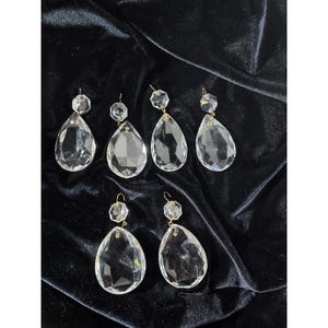 May include: Six clear crystal chandelier pendants with gold-colored metal accents. Each pendant features a faceted teardrop shape below a smaller, octagonal crystal. The pendants are displayed against a dark, velvety background.