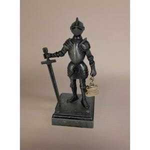 May include: A silver-colored knight figurine holding a sword and a small gold-colored purse. The knight is wearing full armor and a helmet. The purse has the word "ATHY" on it. The figurine is standing on a rectangular base.