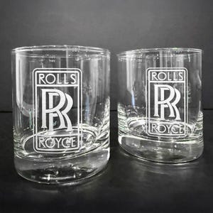 Puede incluir: Dos vasos de vidrio transparentes con el logotipo de Rolls Royce grabado en la parte delantera. El logotipo es blanco y presenta la inscripción Rolls Royce y el icónico emblema doble R.
