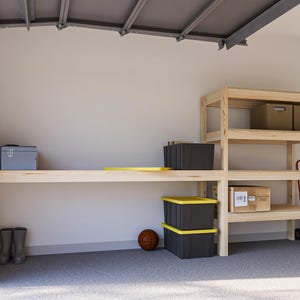 May include: A wooden workbench with a shelf above it. The workbench is made of unfinished wood and has a light brown color. There are several storage bins and boxes on the shelves, as well as a basketball.
