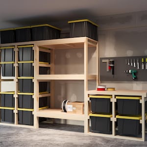 May include: A wooden storage shelf unit with multiple shelves and black storage bins. The unit is in a garage setting with a pegboard wall and a workbench with additional storage bins.