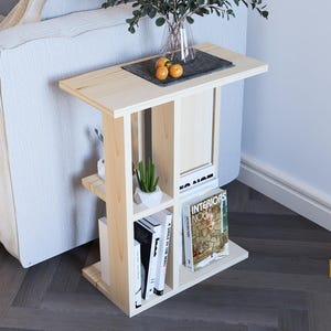 May include: A light wood side table with a shelf and open compartments. The table has a natural wood finish and is decorated with a small potted plant, a tray with oranges, and books with titles like "Interiors Now" and "A Smile in the World".