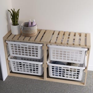 May include: A wooden laundry sorter with two tiers of white plastic baskets. The top shelf holds a woven basket, a potted plant, and bottles. The sorter is made of light-colored wood and is placed against a white wall.