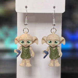 May include: A pair of dangle earrings featuring miniature figurines. Each earring depicts a character with large green eyes, a green tunic, and a blue accessory. The figurines are attached to silver-colored fish hook ear wires.
