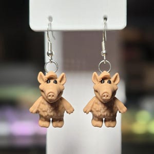 May include: A pair of novelty earrings featuring small, tan-colored figures with large ears and a distinctive snout. The figures are suspended from silver-colored fish hook ear wires. The earrings are displayed against a white background.