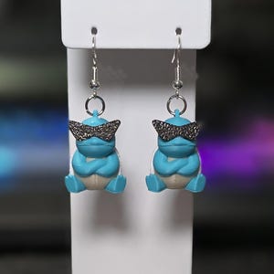 May include: A pair of novelty earrings featuring blue cartoon characters with silver-colored sunglasses. The earrings have silver-colored hooks and are displayed on a white card.