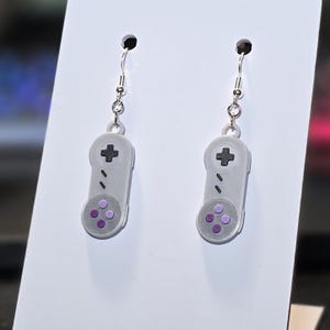 May include: A pair of gray earrings shaped like retro video game controllers. Each earring features a black directional pad, two black buttons, and four purple buttons. The earrings hang from silver-colored hooks.