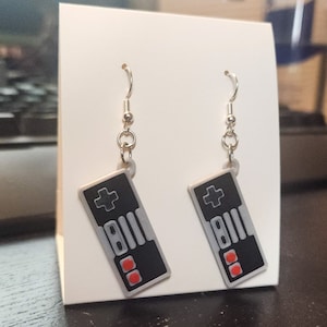 May include: A pair of silver earrings with black and gray Nintendo controller charms. The controllers have red buttons and a white plus sign.