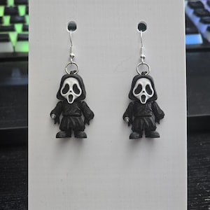 May include: A pair of novelty earrings featuring small, black figures with white, screaming mask faces. The earrings hang from silver-colored fish hook ear wires. The figures are displayed on a white card.