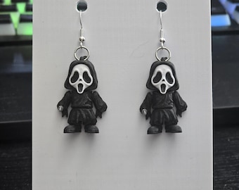 Op de afbeelding: Een paar novelty oorbellen met kleine, zwarte figuren met witte, schreeuwende maskergezichten. De oorbellen hangen aan zilverkleurige oorhaken. De figuren worden gepresenteerd op een witte kaart.
