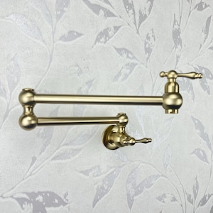 Puede incluir: Un grifo de cocina dorado con un caño giratorio y dos manijas. El grifo está montado en una pared.