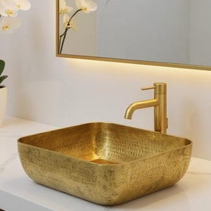 May include: A contemporary bathroom scene featuring a gold square sink and tap. A white countertop holds a stack of white towels and a potted orchid. A gold-framed mirror is above the sink, and the walls are white with grey veining.